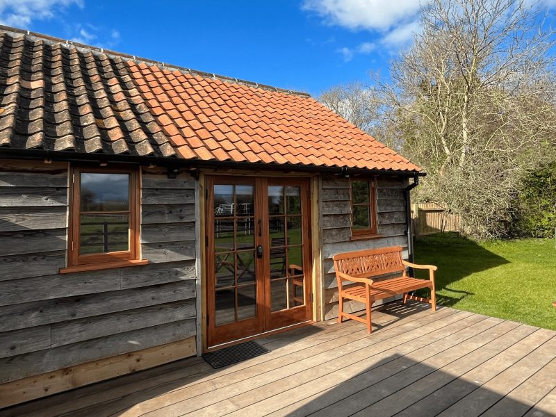 Timber-clad stable conversion with a red pantile roof, glazed double doors and a sunny deck with a wooden bench at Cocklebury Farm, Wiltshire