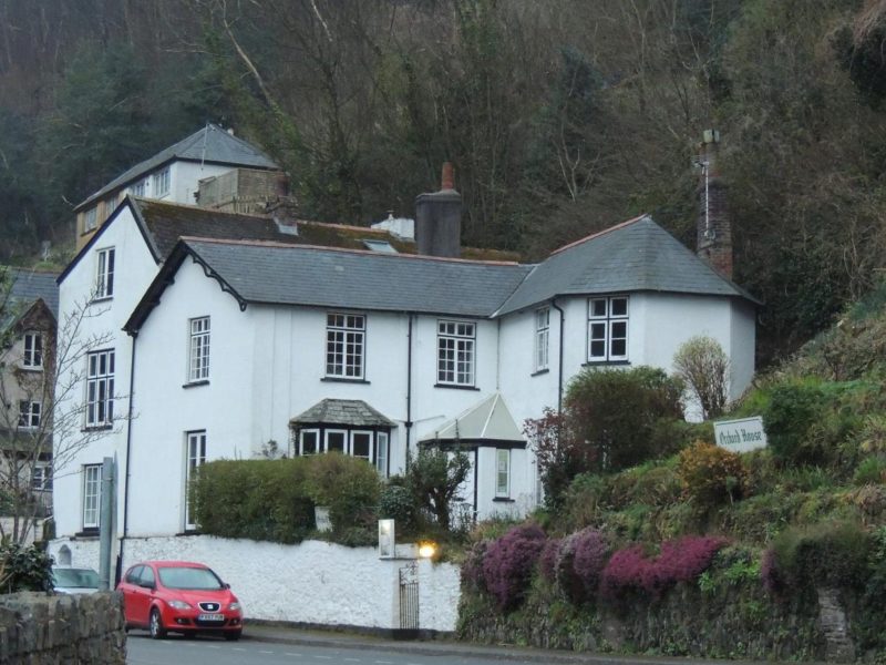 Orchard House Hotel seen from Watersmeet Road in Lynmouth, a white-painted house with bay windows and a heather-filled bank below the wooded hillside