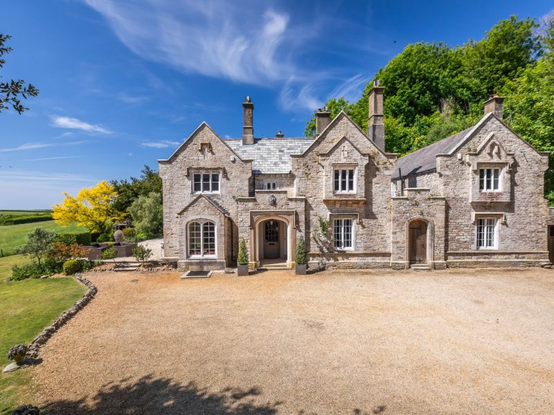 Front of Upwey House, a gabled Grade II-listed stone manor near Weymouth, Dorset, with a gravel drive under blue skies