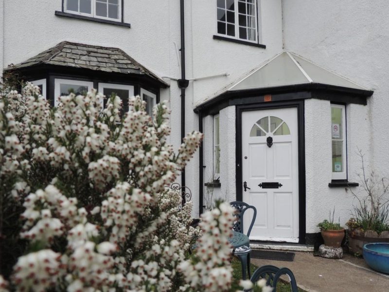 Front door of Orchard House Hotel between two bay windows, with white heather in flower in the front garden