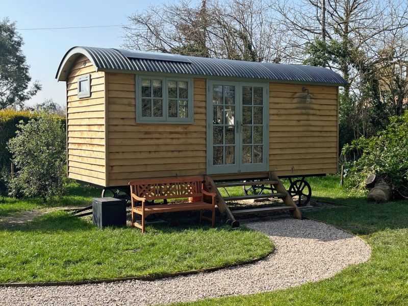 Timber shepherd’s hut with a curved tin roof on cast-iron wheels, reached by a gravel path across the lawn at Cocklebury Farm