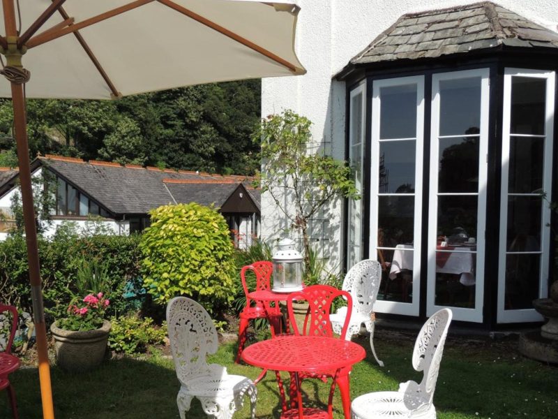 Garden at Orchard House Hotel with a red bistro table, white wrought-iron chairs and a parasol beside a glazed bay window