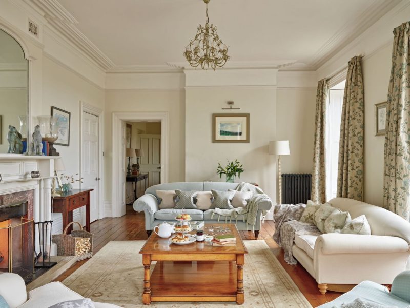 Drawing room at Upwey House with cream sofas, an open fireplace, a chandelier and tall windows onto the garden