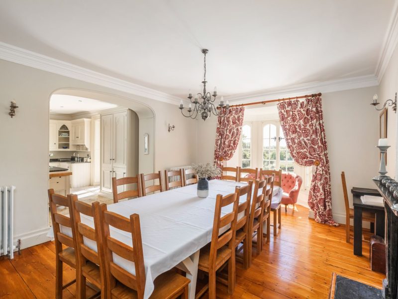 Dining room at Upwey House with a long oak table, chandelier and red patterned curtains, opening into the kitchen
