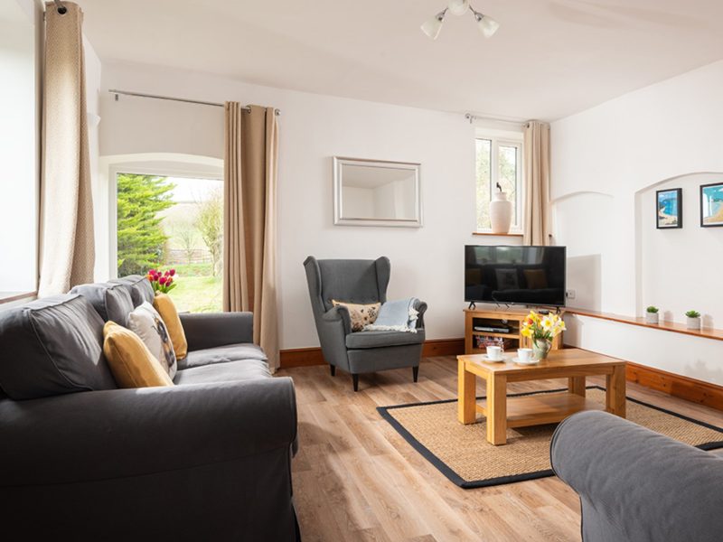 Living room at Mullen cottage, Pitt Farm, with grey sofas, an armchair, wooden coffee table and garden doors